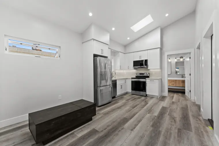 Open living area with vaulted ceiling, skylight, and wood-look plank floor leading to a white kitchen with stainless appliances