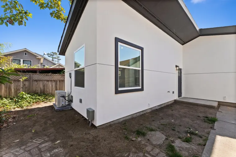 Side corner of the ADU with a black-trimmed window, the AC condenser, and a brick-edged yard along the fence