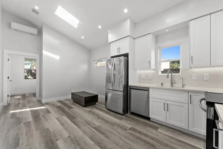 Great room under a vaulted, skylit ceiling, opening to a white shaker kitchen with stainless appliances