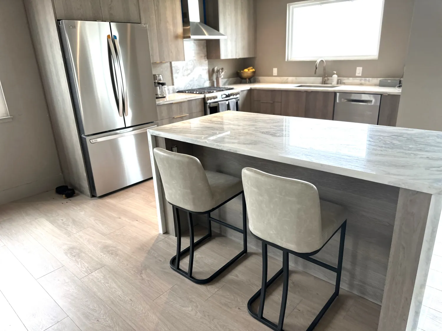 Kitchen with gray wood-grain flat-panel cabinets, a quartzite waterfall island with two upholstered stools, stainless refrigerator, and light plank floors