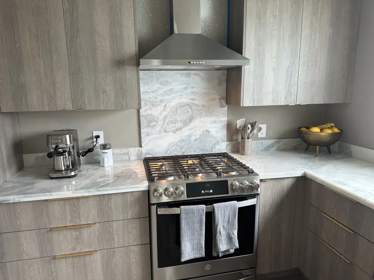 Stainless gas range under a chimney hood with a full-height veined stone backsplash, gray wood-grain cabinets, and brass pulls