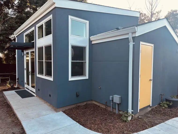 Blue-gray stucco ADU with white trim, tall corner windows, French doors under a metal awning, and a yellow side door along a curved concrete path