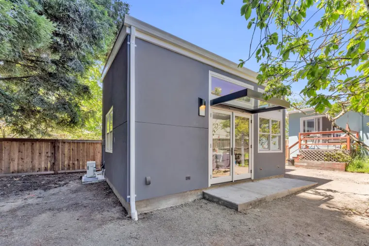 Blue-gray ADU with a shed roof, clerestory windows, and French doors opening onto a concrete pad, framed by trees and a wood fence
