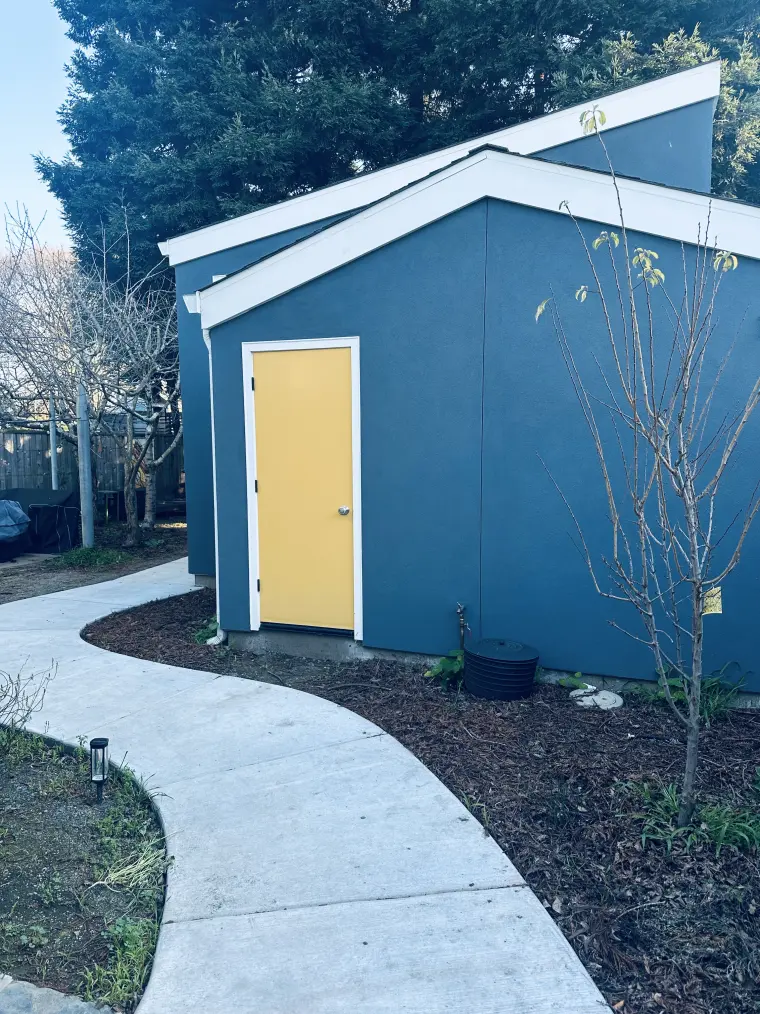 Curved concrete path through mulched beds leading to the ADU's yellow side door and white-trimmed gable