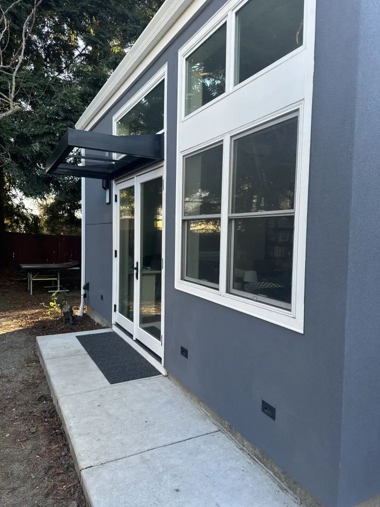 Side of the ADU with stacked white-trimmed windows and French doors under a metal awning, with a door mat on a concrete pad
