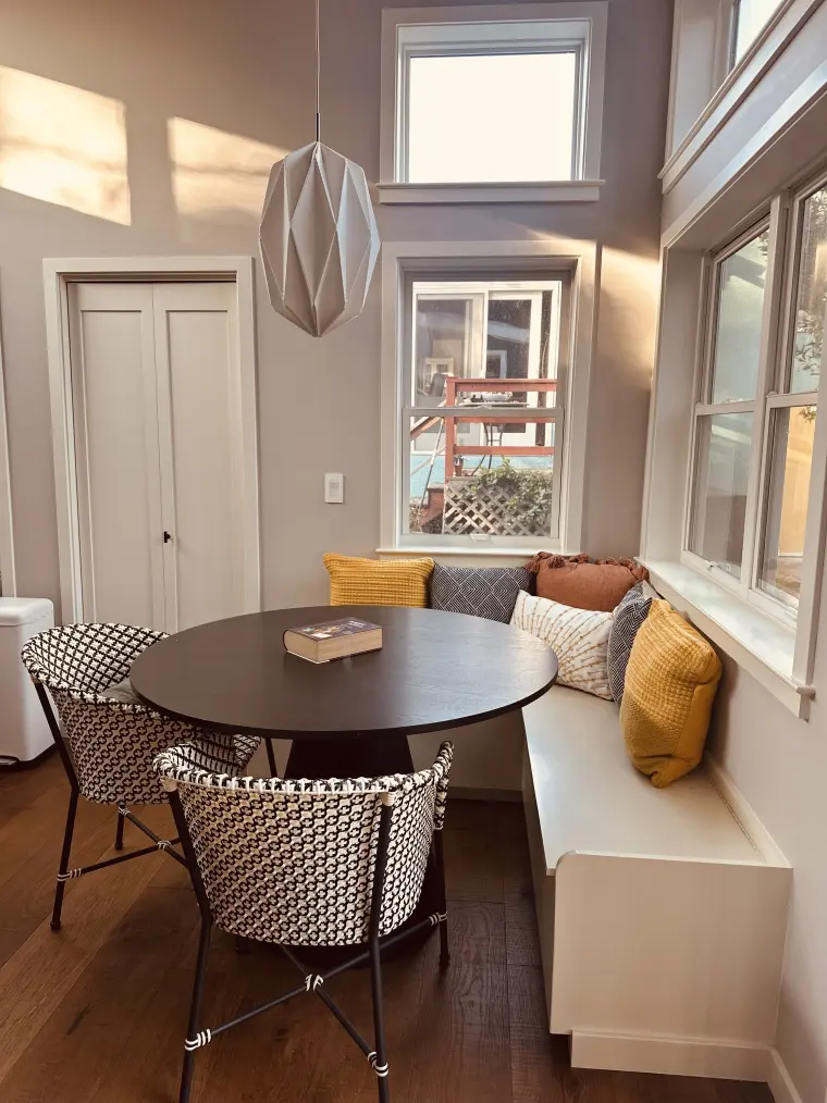 Built-in bench dining nook with round black table, woven chairs, throw pillows, and a folded paper pendant light under tall windows