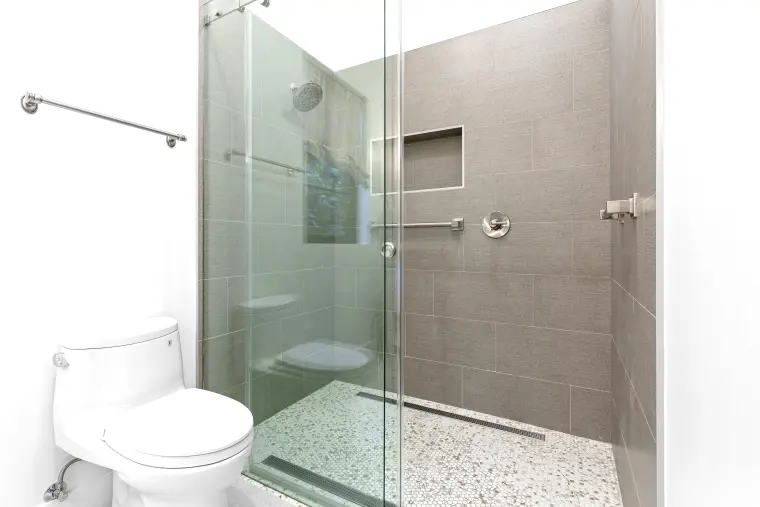 Walk-in shower with frameless glass, large gray wall tile, recessed niche, and speckled floor tile beside a white toilet