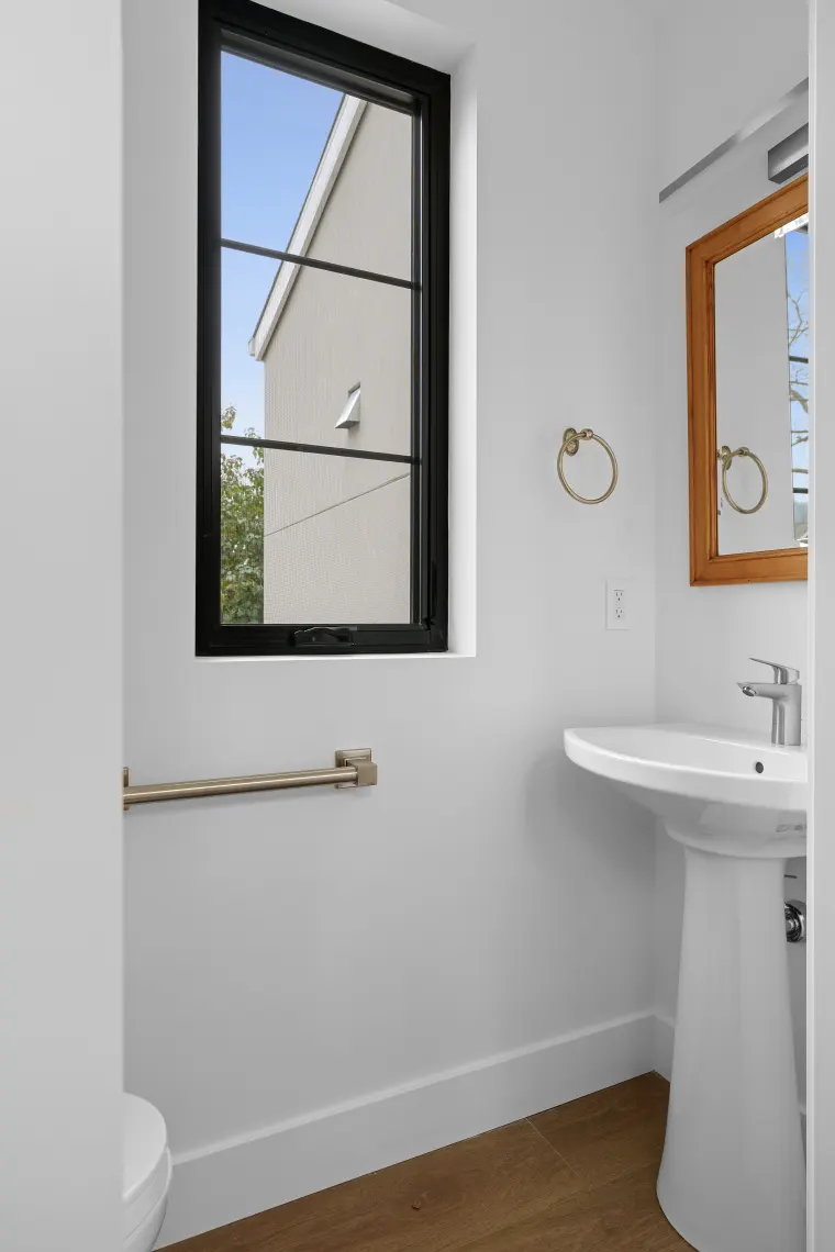 Powder room with pedestal sink, wood-framed mirror, black-framed window, and brushed grab bar