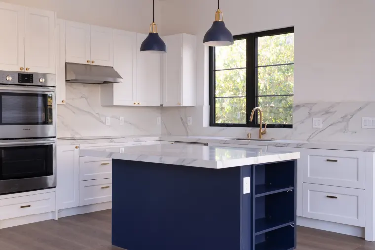 Navy kitchen island with white shaker cabinets, double wall ovens, and a marble-look backsplash