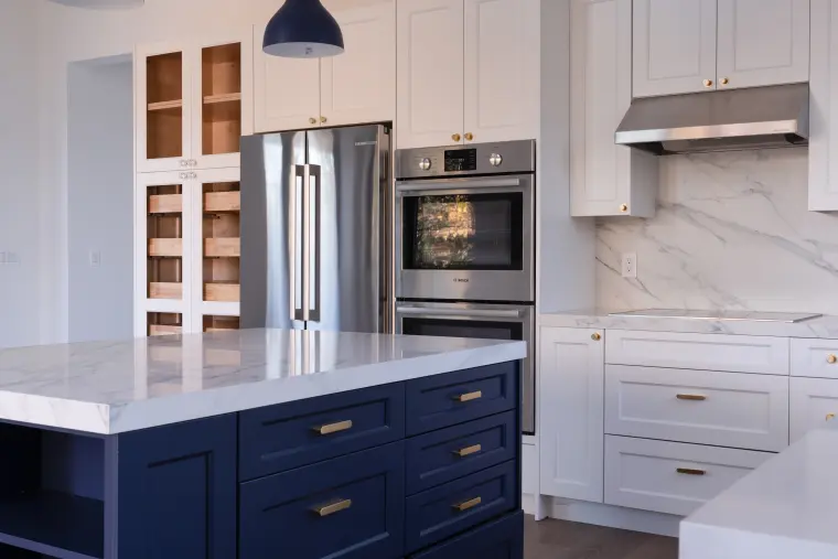 White shaker kitchen with a stainless refrigerator, wall ovens, and a navy island with brass pulls
