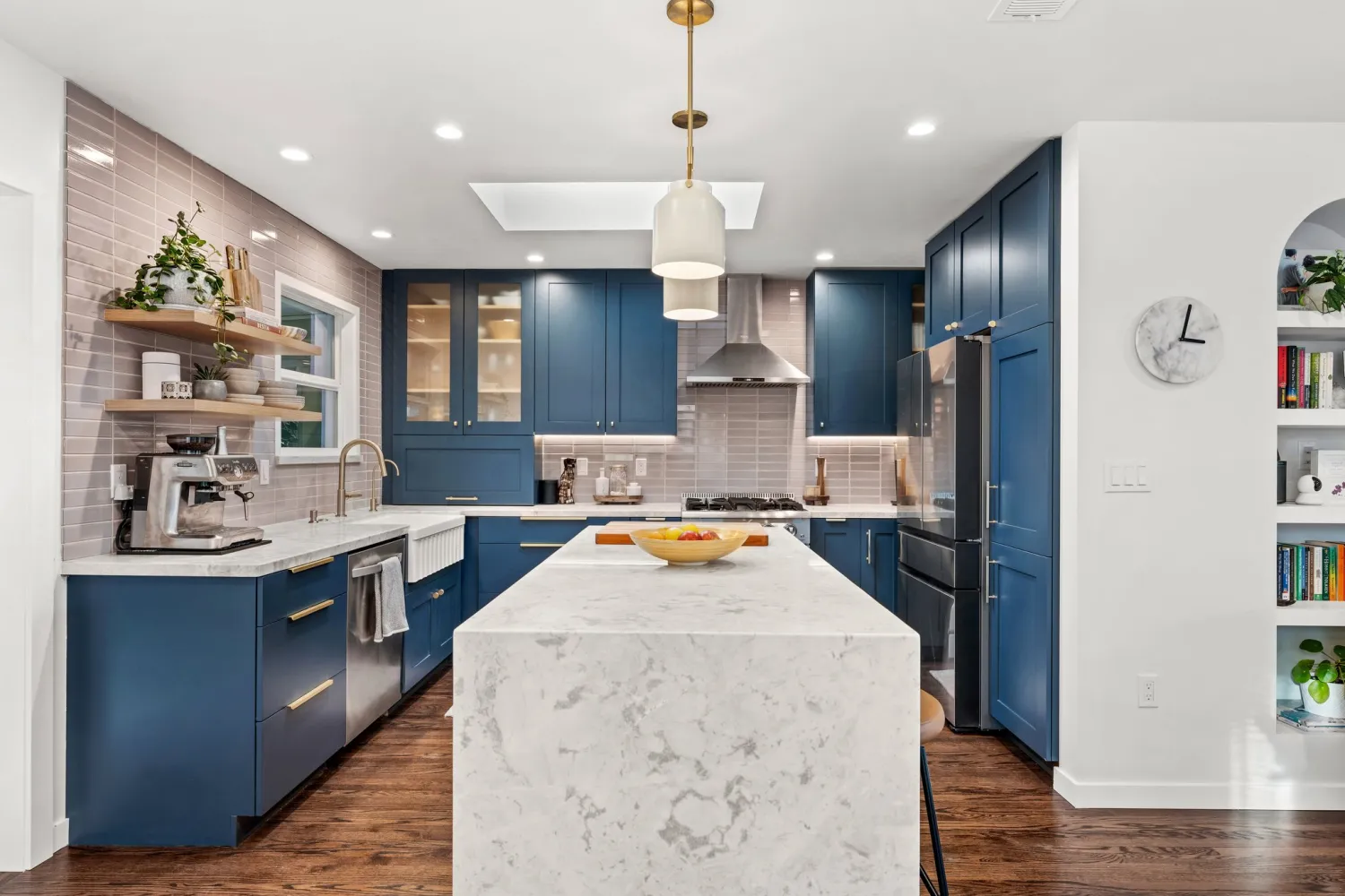 Kitchen with navy shaker cabinets, a white quartz waterfall island, gray stacked tile backsplash, floating wood shelves, and a skylight