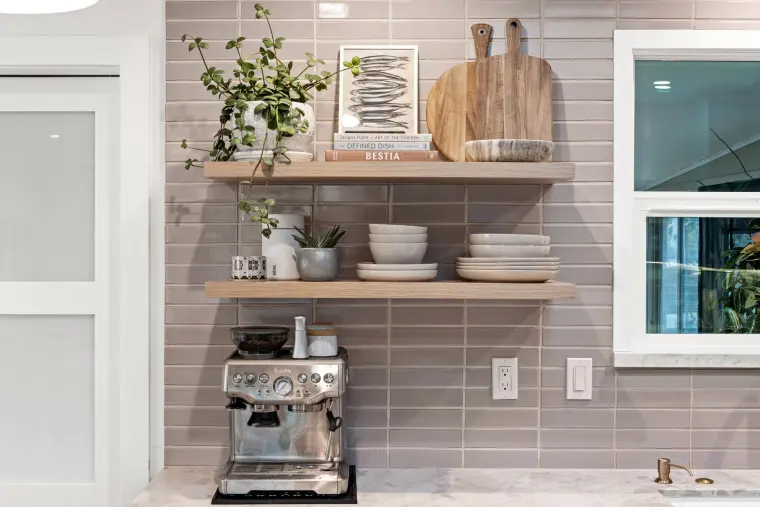 Floating wood shelves with dishes and cutting boards above an espresso machine on a gray stacked tile backsplash