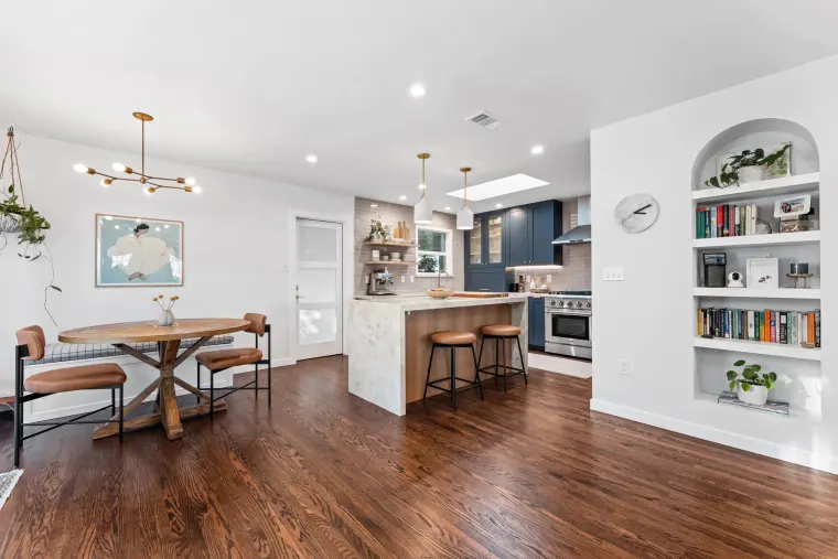 Open plan with a round dining table under a brass chandelier, kitchen island with stools beyond, and an arched built-in bookshelf
