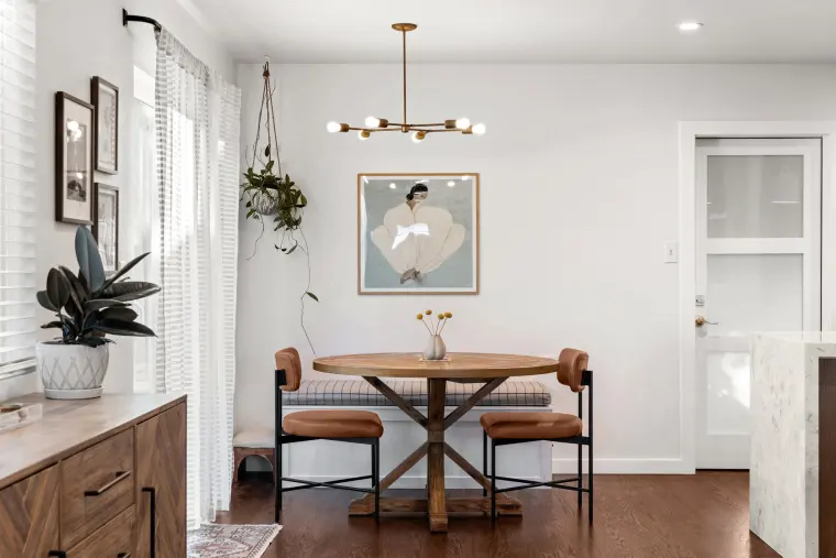 Dining nook with a round wood pedestal table, cognac leather chairs, brass chandelier, and a framed print on white walls