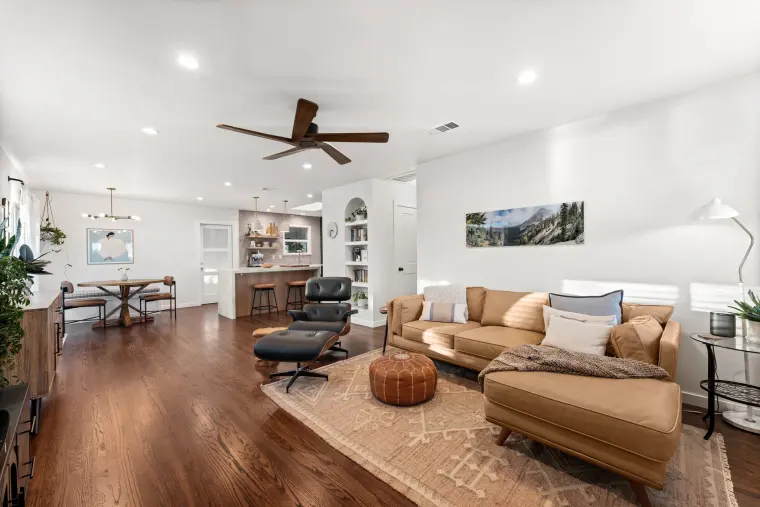 Living room with a tan sectional, Eames-style lounge chair, dark hardwood floors, and a ceiling fan, open to dining and kitchen