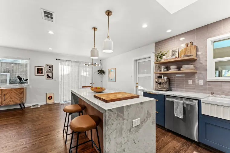 Island with wood cutting board and stools beside a run of navy base cabinets, stainless dishwasher, and apron-front sink under floating shelves