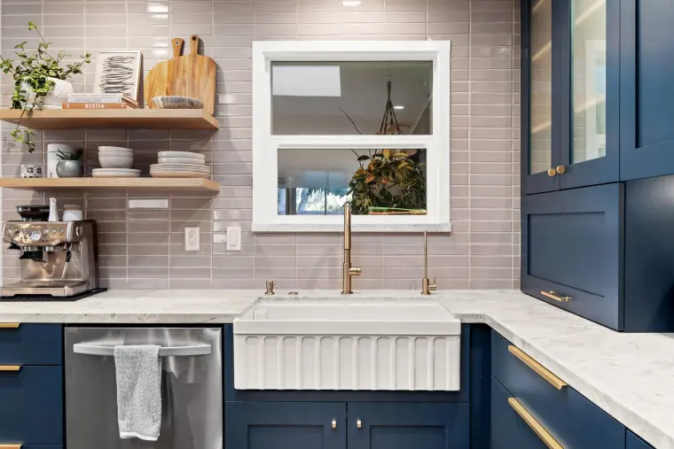 White fluted apron-front sink with brass faucet under a window, gray stacked tile, and floating wood shelves with dishes