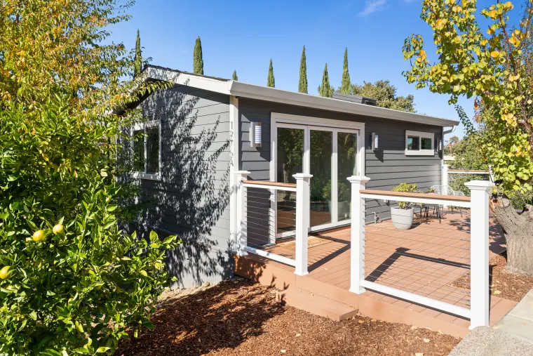 Gray lap-sided ADU with glass doors and a white cable-rail deck, seen past a citrus shrub under blue sky