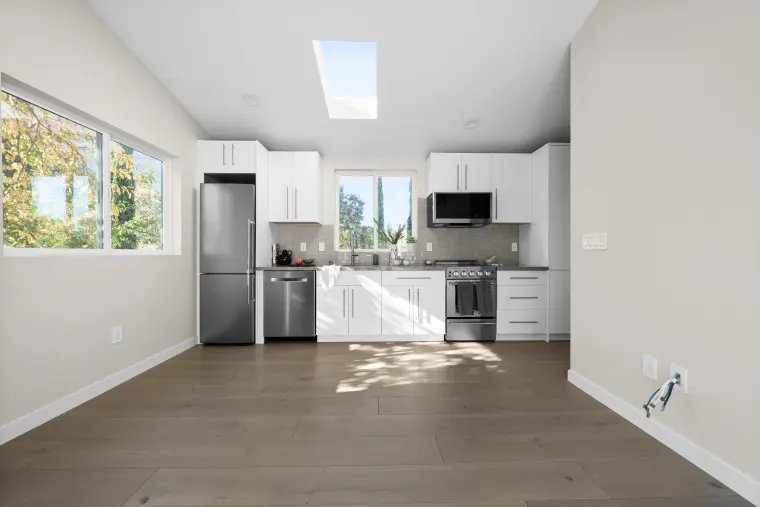 Open room looking toward a white kitchen with stainless appliances, a skylight, and wide-plank floors
