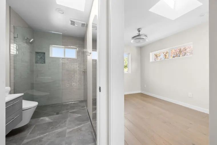 View from the hall into a gray-tiled glass shower on one side and a bedroom with clerestory windows on the other