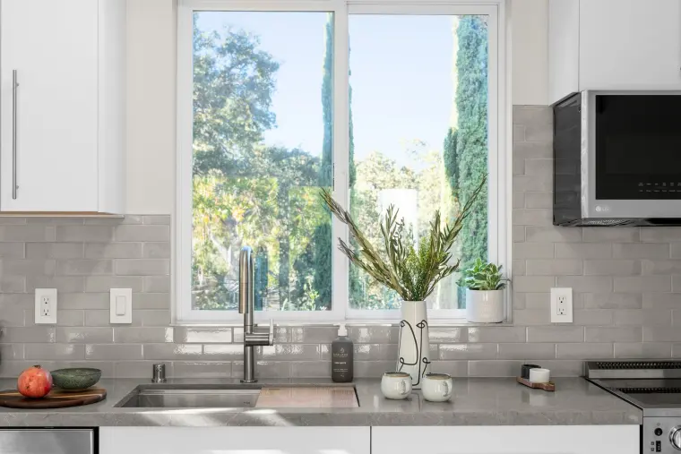 Kitchen sink under a window framing trees, with gray subway tile and quartz counters