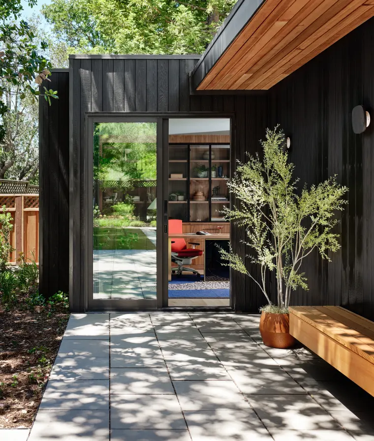 Paver path between charred-cedar walls under a cedar overhang, with a glass door, potted tree, and wood bench