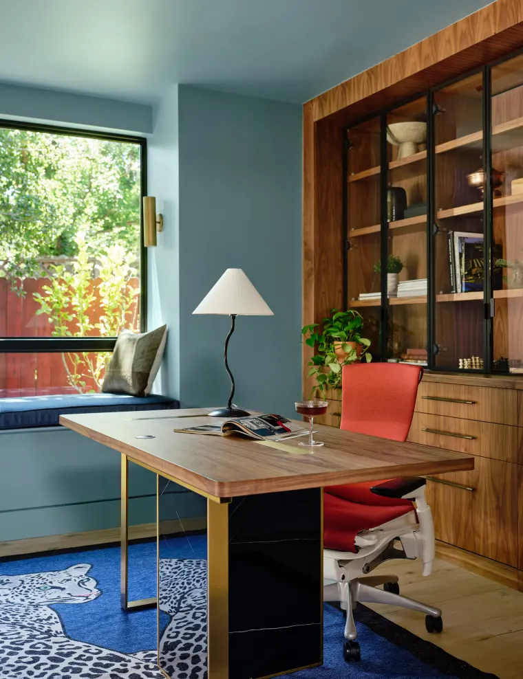 Home office with a wood desk, red chair, and built-in walnut shelving beside a window seat