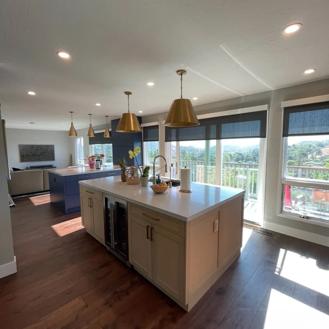 Wide view of the finished open kitchen with pale wood and navy islands, brass pendant lighting, living room with gray sofa in the background, wall of windows showing sweeping hillside and valley views