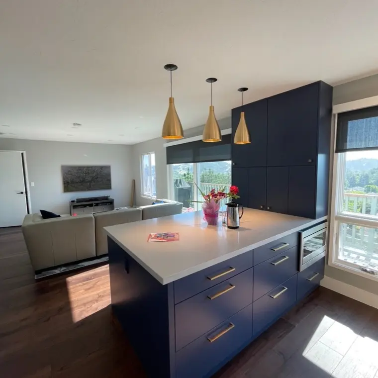 Finished navy blue kitchen island with brass pendant lighting opening onto an adjoining living room with gray sectional sofa, framed orchard artwork, and large windows with hillside views