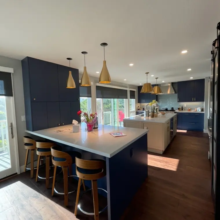 Finished open kitchen with navy blue cabinetry, white quartz waterfall island, brass conical pendant lights over a bar-seating peninsula, second island with pale wood cabinetry, dark hardwood floors, deck door with hillside view