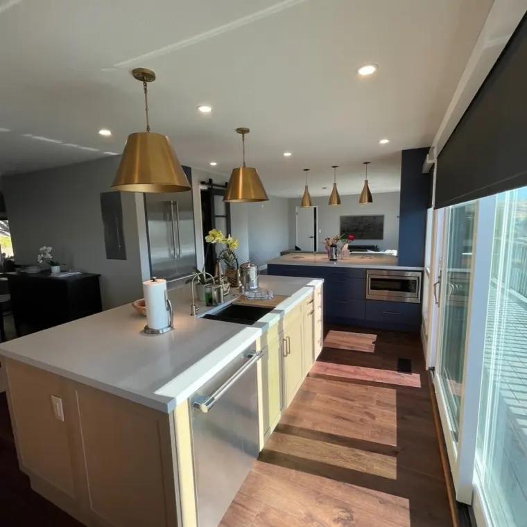 Finished kitchen with two islands (pale wood with wine fridge, navy with microwave drawer), oversized brass dome pendants, dark hardwood floors, dining nook with three matching pendants and orchard artwork in the background