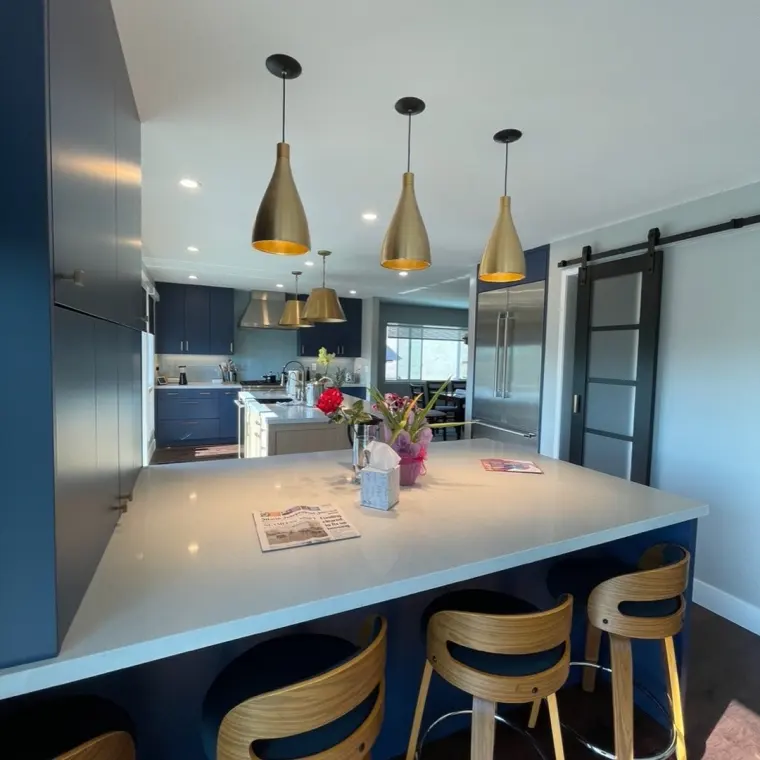 View across the finished kitchen island toward the range wall and a black-framed barn door, brass pendant lights, stainless refrigerator, navy cabinetry, dining table glimpsed through the pass-through