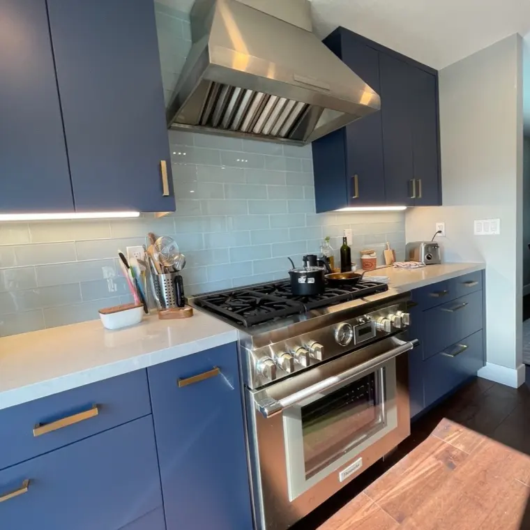 Finished cooktop wall, navy cabinetry with brass pulls, light blue subway tile backsplash, professional range with stainless hood, toaster and utensil crock on the counter, walnut cutting board on the island edge
