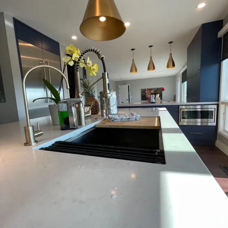 Close-up of the finished prep-sink island with gooseheck brass faucet and pot filler, black integrated sink drainboard, quartz waterfall counter, orchid on the ledge, dining pendants and stainless microwave drawer in the background