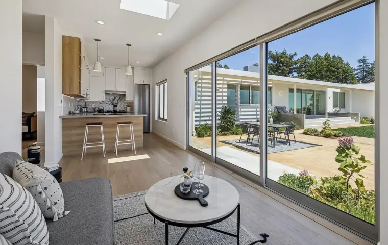 Living area with a corner of wall-to-wall sliding glass looking onto the patio, round coffee table, and the kitchen beyond