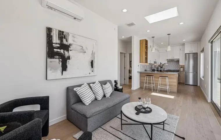 Open living area with gray sofa, round coffee table, and abstract art, with a kitchen island and bar stools beyond