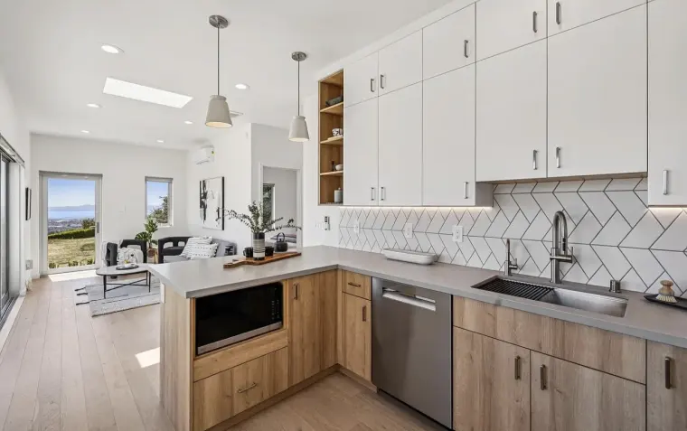 Kitchen with wood lower cabinets, gray counters, white herringbone backsplash, and pendant lights, opening to the living area