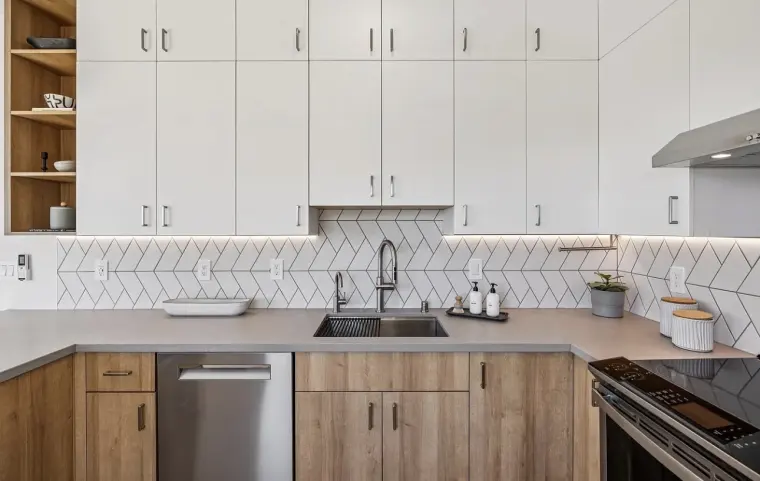 Sink wall with wood lower cabinets, gray counters, white herringbone tile backsplash, and stainless dishwasher