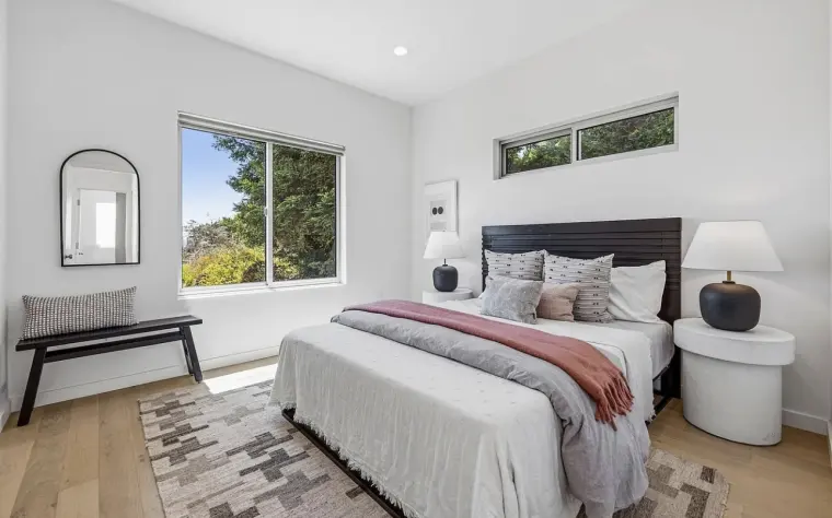 Bedroom with an upholstered bed, bench, arched mirror, and windows looking onto trees, on light oak flooring