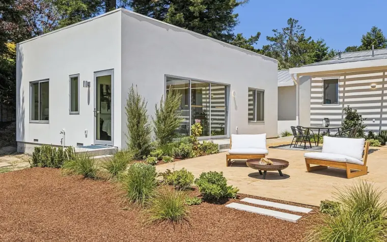 White stucco ADU with a sliding glass door and aluminum windows beside a decomposed-granite patio with lounge chairs, a fire bowl, and new plantings