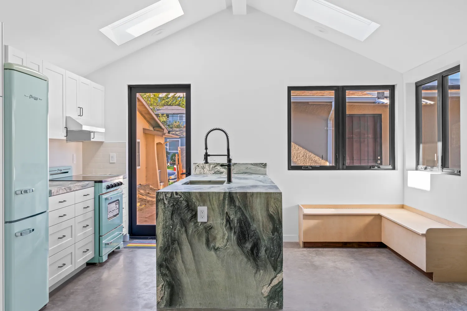 Vaulted kitchen with skylights, a waterfall-edge stone island, mint retro refrigerator and range, and a built-in bench under black-framed windows
