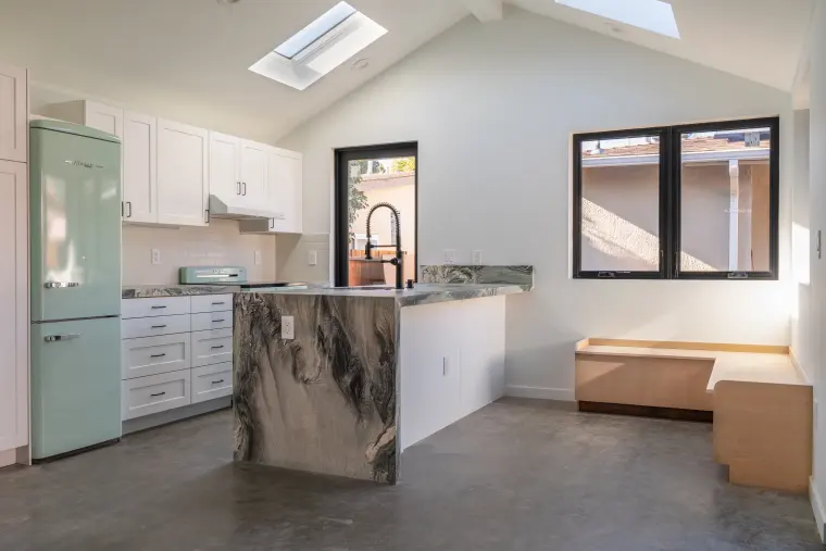 Angled view of the vaulted kitchen with stone waterfall island, white shaker cabinets, mint appliances, and a wood bench below corner windows
