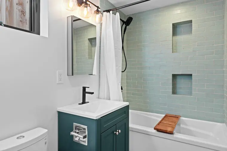 Finished ADU bathroom with sage-green subway-tile tub surround with tiled niches, dark teal vanity, wood bath tray
