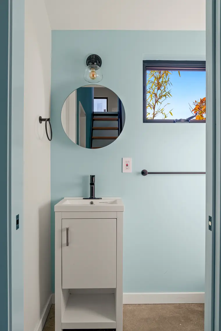 Powder room with pale aqua walls, round mirror, small white vanity, black towel bar, and a window