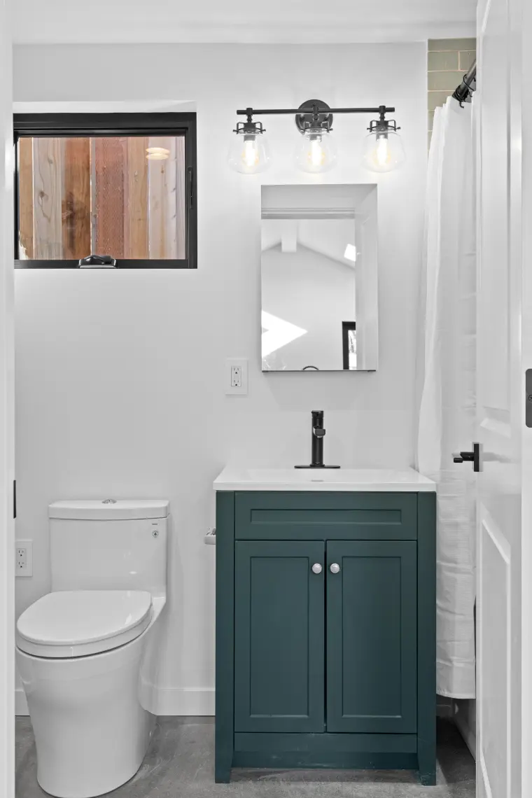 Bathroom with dark green shaker vanity, three-light fixture, mirror cabinet, window, and toilet on a concrete floor