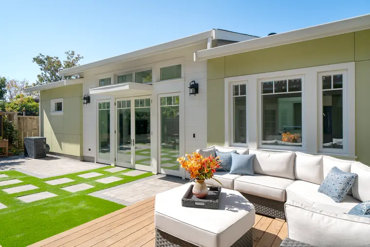 Sage-green and white ADU with shed roofs, clerestory windows, and French doors, seen from a deck with an outdoor sectional