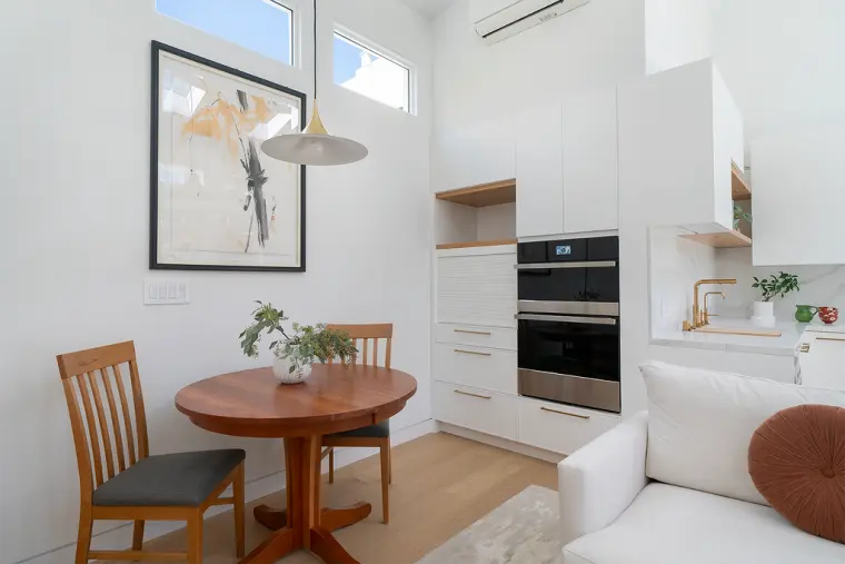 Dining spot with a round wood table under a brass pendant, beside a white kitchen wall with a double wall oven