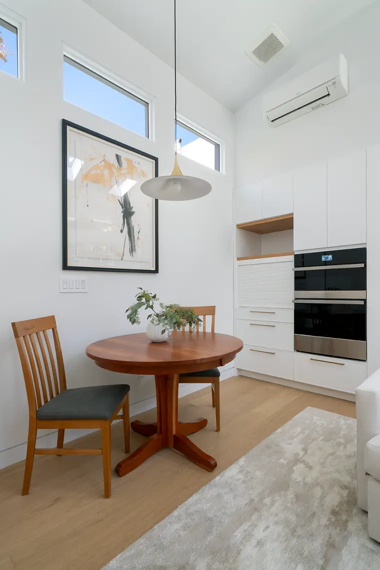 Round dining table under a brass pendant and clerestory windows, with the kitchen’s wall oven alongside
