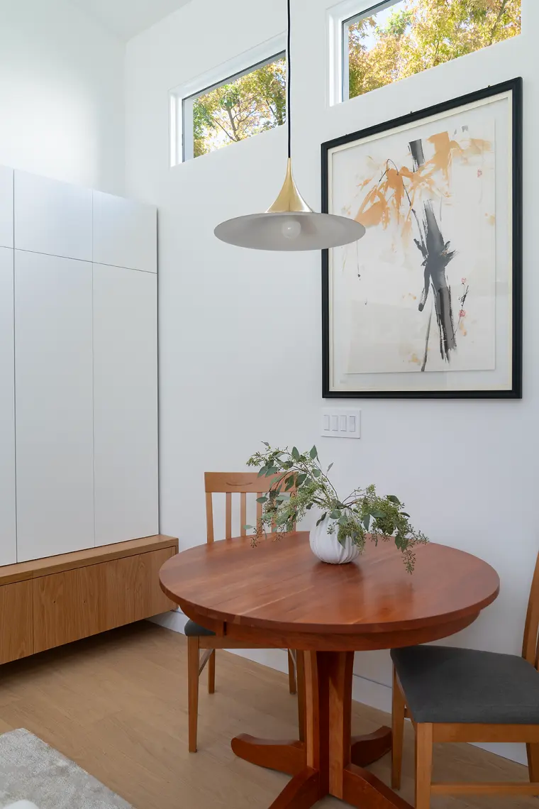 Dining corner with a round wood table, a brass pendant, framed art, and clerestory windows