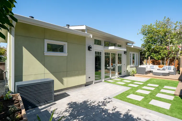 Side of the ADU in sage-green panels with a small window, the heat pump screened behind a slatted cover, and the patio beyond